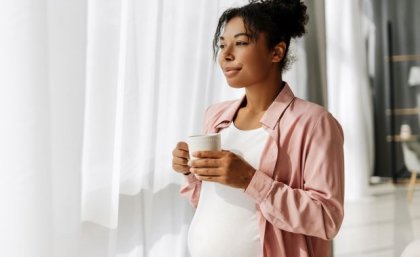 A pregnant woman holds a coffee mug and looks through sheer curtains out a window. 